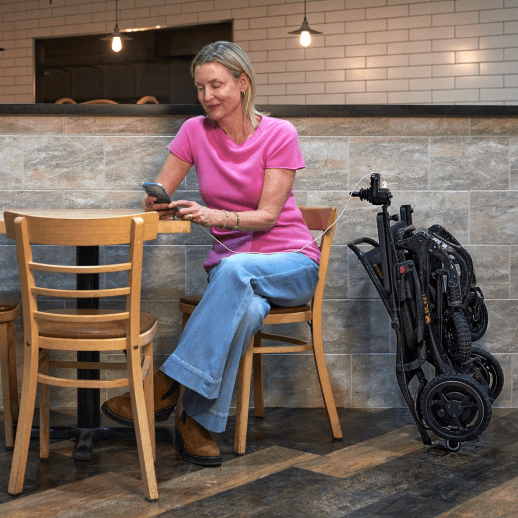 A woman in a pink top and blue jeans sits at a café table using her phone. Next to her, the lightweight Cricket – Power Wheelchair by Golden is folded and charging amidst cozy wooden chairs and tiled walls.