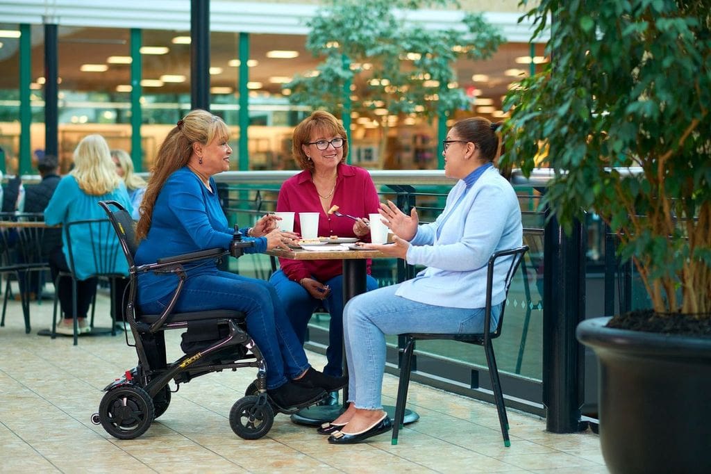 Three women sit around a table in a bright indoor café, chatting and smiling. One woman uses the Cricket - Power Wheelchair by Golden. They enjoy drinks and snacks, with greenery and other people in the background.