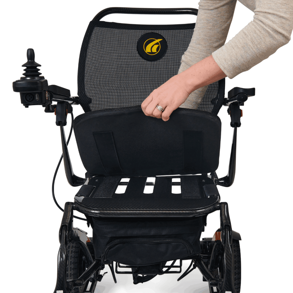 A person lifts the seat cushion of a black Cricket - Power Wheelchair by Golden, which features a lightweight frame, mesh backrest, and joystick control, revealing the space beneath the seat.