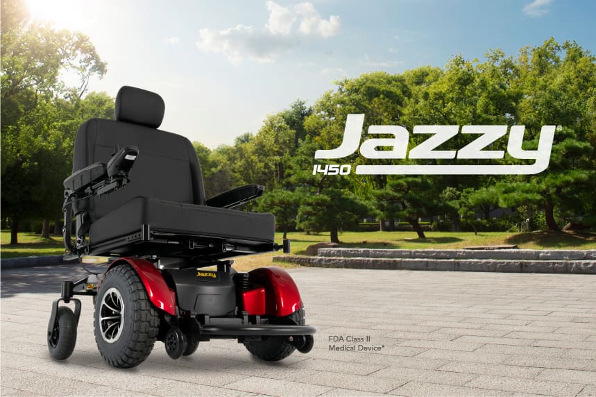 A black and red Jazzy 1450 - Power Wheelchair by Pride Mobility is parked on a sunny paved path, with green trees and blue sky behind it. The product name appears prominently in the image.