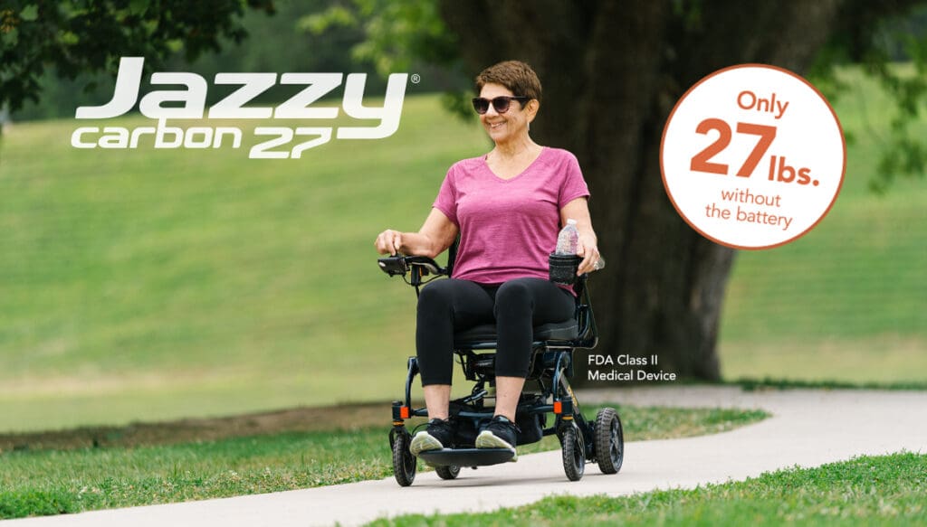 A smiling woman rides the Jazzy Carbon 27- Power Wheelchair by Pride Mobility on a park path. Text highlights this lightweight wheelchair weighs just 27 lbs (no battery) and is an FDA Class II medical device.