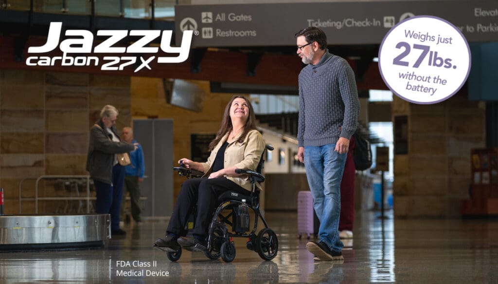 A woman in a portable Jazzy Carbon 27X Power Wheelchair by Pride Mobility smiles at a man walking beside her in an airport. Text reads: Weighs just 27 lbs. without the battery; FDA Class II Medical Device.
