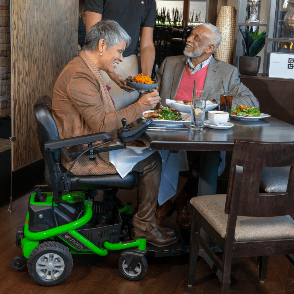 Two older adults smile and enjoy a meal at a restaurant. One uses the LiteRider Envy - Power Wheelchair by Golden as a server hands them a bowl of food.