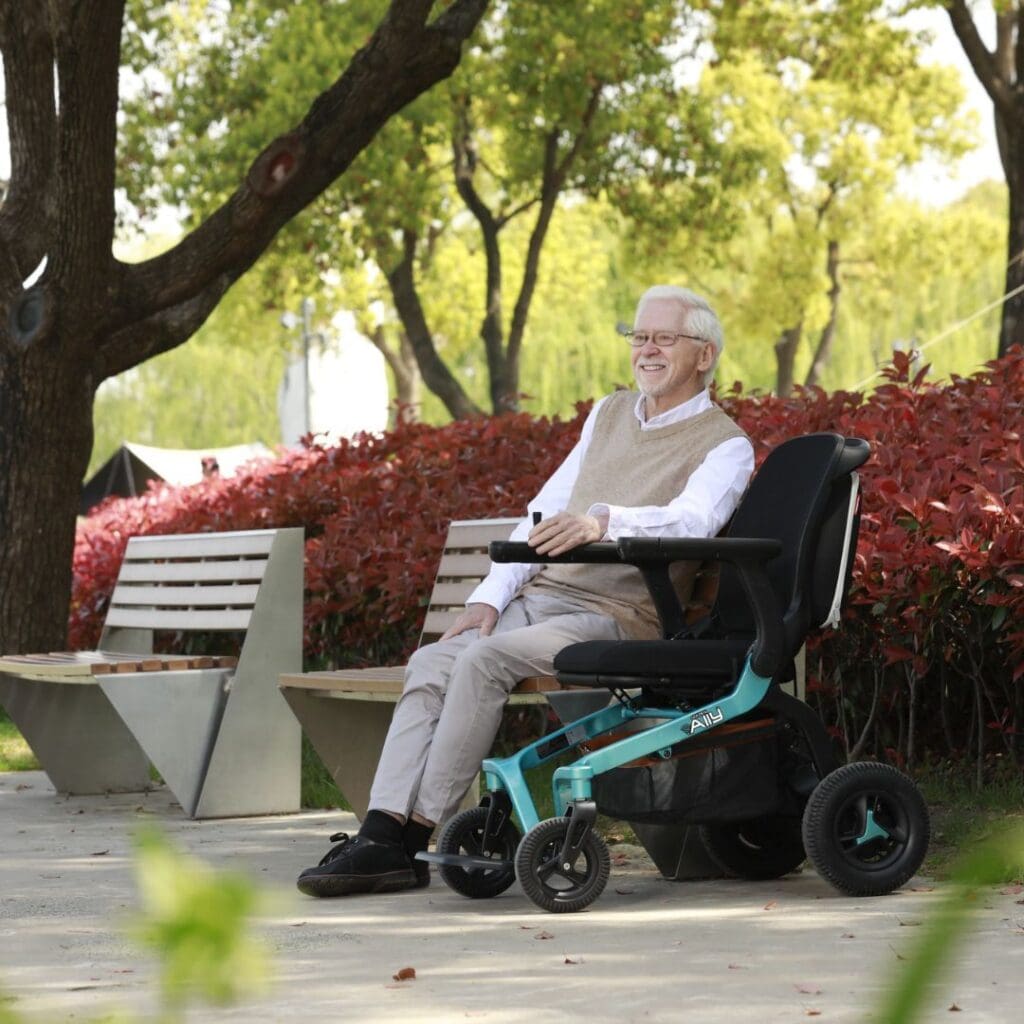 An older man with white hair and glasses smiles while sitting in the Ally Power Wheelchair by Golden on a sunny park path, surrounded by trees, red shrubs, and benches.