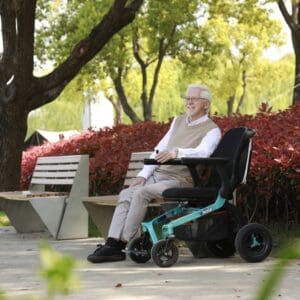 An older man with white hair and glasses smiles while sitting in the Ally Power Wheelchair by Golden on a sunny park path, surrounded by trees, red shrubs, and benches.