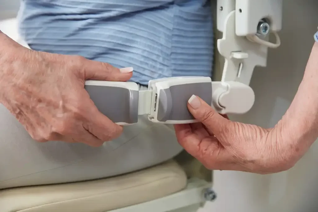 Close-up of an elderly person securing a safety belt on the Handicare 1100 Straight Stair Lift by Handicare, highlighting their hands and part of the stair lift mechanism.