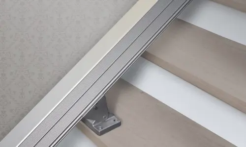 Close-up of a Handicare 1100 Straight Stair Lift rail mounted along a staircase with light-colored steps and patterned wallpaper in the background.