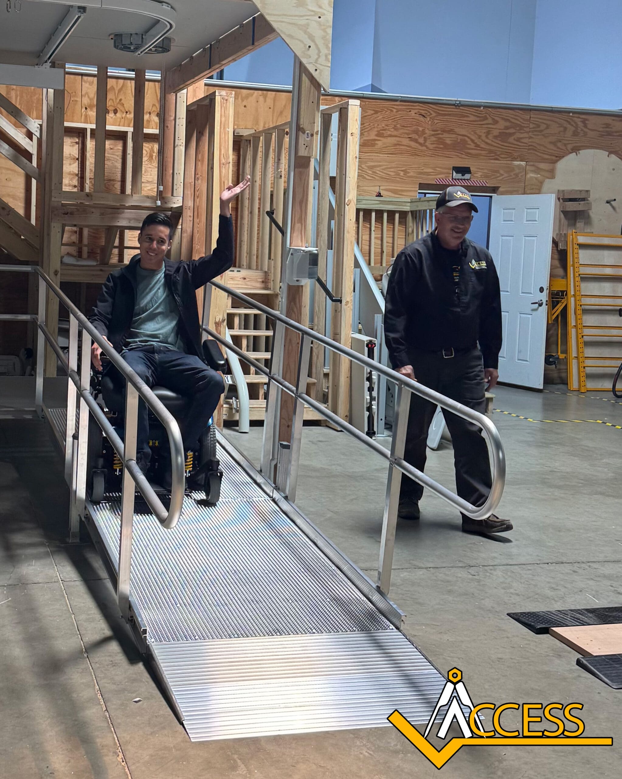 A smiling person in a wheelchair raises their hand while using an indoor metal ramp, accompanied by another person standing beside the ramp. Wooden structures and ACCESS logo are visible in the background.