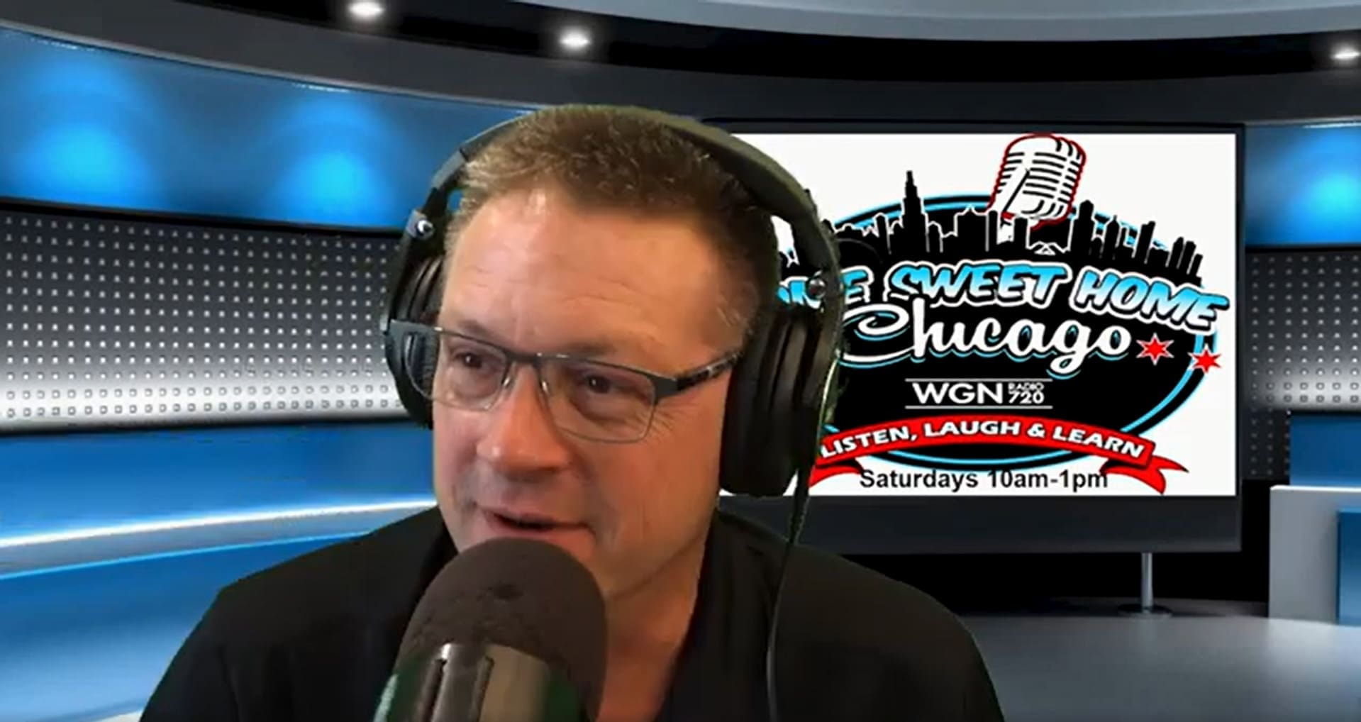 A man wearing glasses and headphones speaks into a microphone in a studio. Behind him, a screen displays the Home Sweet Home Chicago WGN radio show logo, schedule information, and highlights topics like grab bars for bathroom safety and accessibility.