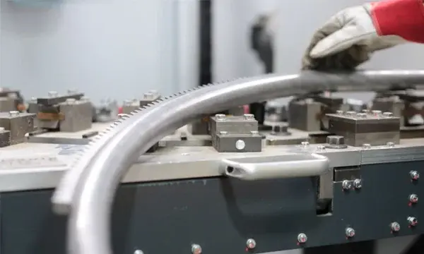 A gloved hand adjusts a large, curved metal pipe on an industrial bending machine, showcasing the precision engineering found in manufacturing the Handicare 4000 Curved Stair Lift by Handicare.