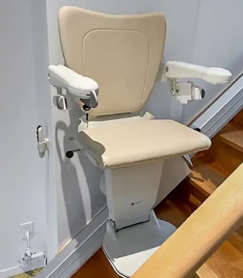 The Handicare 1100 Straight Stair Lift by Handicare is installed along a wooden staircase, featuring a beige seat and wall-mounted rail to assist individuals with mobility challenges in residential settings. The Handicare 1100 Straight Stair Lift by Handicare is installed along a wooden staircase, featuring a beige seat and wall-mounted rail to assist individuals with mobility challenges in residential settings.
