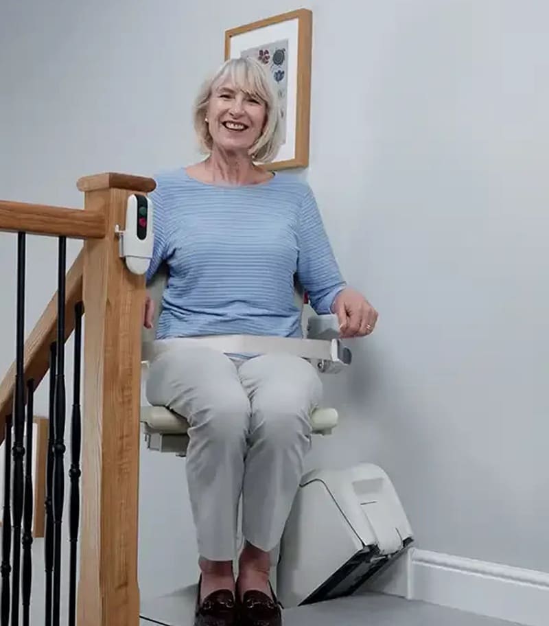 A smiling older woman with gray hair, in a light blue top and beige pants, sits at the bottom of her stairs on a Handicare 1100 Straight Stair Lift by Handicare. A small framed picture is visible on the wall behind her. A smiling older woman with gray hair, in a light blue top and beige pants, sits at the bottom of her stairs on a Handicare 1100 Straight Stair Lift by Handicare. A small framed picture is visible on the wall behind her.