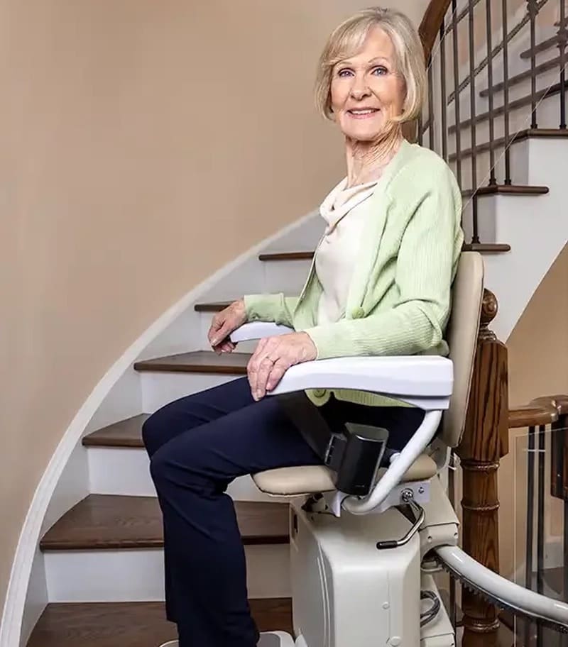 An older woman in a light green cardigan smiles at the camera while seated on a Handicare Stairfriend 23 Curved Stair Lift at the bottom of a curved staircase inside a bright, well-lit home.