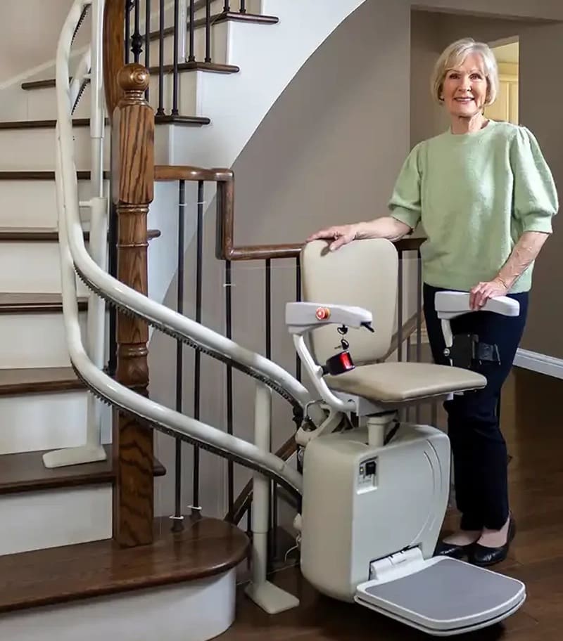 An older woman smiles beside a Stairfriend 23 Curved Stair Lift by Handicare at the base of a wooden staircase in a home, demonstrating the accessibility device.