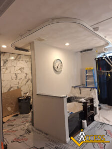 A room under renovation with a partially finished ceiling, exposed curved track, ladder, covered furniture, wall clock, and marble-patterned wall. Protective sheets cover the floor and surfaces. Access logo in bottom right corner.