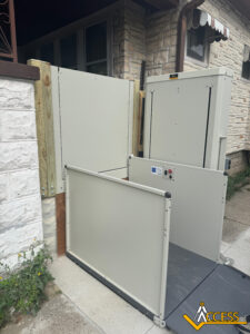 A residential vertical wheelchair lift installed outdoors next to a house, allowing accessible entry. The lift is light-colored with safety rails and control buttons. ACCESS logo is visible in the bottom right corner.