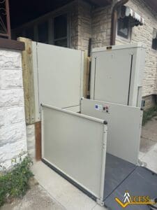 A residential wheelchair lift is installed outdoors beside a house, providing accessible entry. The lift is at ground level, surrounded by white panels, with control buttons visible on one side. The Access logo is in the corner.