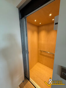 A small, wood-paneled home elevator with a folding metal door is open, revealing a lit interior. A railing is attached to the back wall. The Access company logo is visible in the bottom right corner.