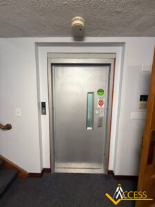 A closed silver elevator door in a building hallway, with a small vertical window, wall buttons, safety and accessibility stickers, and a smoke detector on the ceiling above. ACCESS logo is in the bottom right corner.