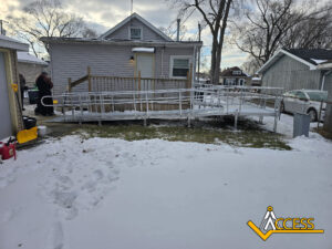 A metal wheelchair ramp leads from a snowy backyard to the door of a small gray house. Two people stand near the entrance. The yard and roofs have snow, and there’s an “ACCESS” logo in the bottom right corner.