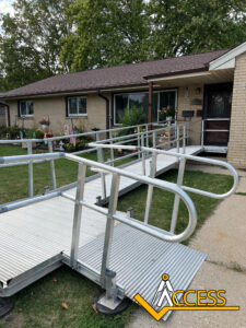 A metal wheelchair ramp with railings leads up to the front door of a single-story brick house. The ACCESS logo appears in the bottom right corner. There are plants and lawn decorations in the yard.