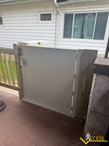 A white wheelchair lift gate installed between a wooden fence and a brick wall on a tiled porch, with a white house and green grass visible in the background. ACCESS company logo is in the bottom right corner.