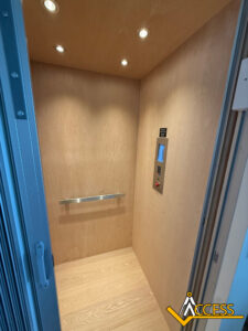 A small, modern wood-paneled elevator interior with a metal handrail, control panel, overhead lights, and an open door. The floor, walls, and ceiling feature light wood. An Access logo appears in the corner.