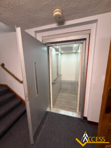 An open elevator with silver walls and light wood flooring is next to a carpeted staircase with a wooden handrail. A smoke detector is visible on the ceiling, and an Access logo is in the corner.