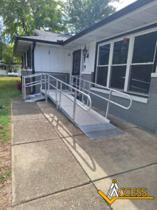 A metal wheelchair ramp with handrails leads to the entrance of a single-story house. The ramp is installed over a concrete walkway. The Access company logo is in the lower right corner.