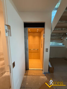 A modern home elevator with wood paneling and metal handrail is open next to a staircase. The ACCESS logo appears in the bottom right corner.