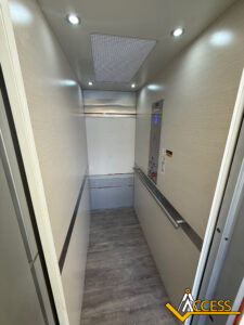 A modern, empty elevator with beige walls, wood-look flooring, metal handrails, and a control panel. The ceiling has recessed lights and a ventilation panel. The Access logo is in the bottom right corner.