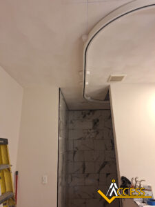 Curved ceiling track with curtain hooks installed above a marble-tiled shower area; a yellow ladder is visible at the left and a vent is on the ceiling. ACCESS logo appears in the lower right corner.