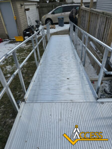 A metal wheelchair ramp leads from a snow-dusted yard to a fenced area near a house. A person stands in the background. The ACCESS logo is visible in the bottom right corner.