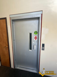 A closed silver elevator door with a small vertical window, caution and fire safety stickers, an Access logo on the wall, and an elevator button panel next to the door.