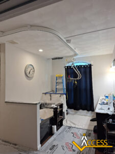 A room under renovation with a covered sink, a step ladder, and a ceiling-mounted track system. Black curtains cover the window, a clock is on the wall, and protective sheets cover the floor and surfaces. ACCESS logo is in the corner.