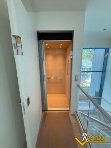 A residential elevator with wood-paneled interior, open accordion-style door, and ceiling lights is located next to a glass railing on an upper floor. The Access company logo is in the bottom right corner.