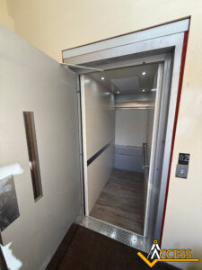 Open elevator with silver walls, wood-patterned floor, and ceiling lights. The elevator door is partially open, showing the interior. The Access logo is visible at the bottom right corner near the control panel.