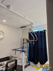 A ceiling track hoist system is installed in a room with a white ceiling, dark blue curtains, a wall clock, and a blue ladder beside covered furniture. The ACCESS logo is visible in the bottom right corner.