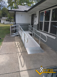 A metal wheelchair ramp with handrails leads to the front door of a house. The ramp is installed over a concrete walkway. Trees and a lawn are visible in the background. An Access logo is in the bottom right corner.