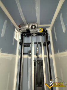 An elevator shaft with a visible motor, cables, and guide rails. The shaft walls are unfinished drywall, and there is an Access company logo in the bottom right corner of the image.