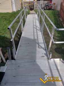 A metal wheelchair ramp with handrails on both sides leads from a house to a fenced yard. The ramp is positioned over grass, and the ACCESS company logo is visible in the corner.