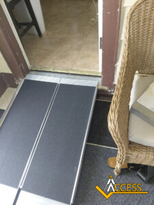 A portable black and silver wheelchair ramp leads over a threshold into a tiled room, next to a wicker chair and a mat with the yellow ACCESS logo at the bottom right corner.