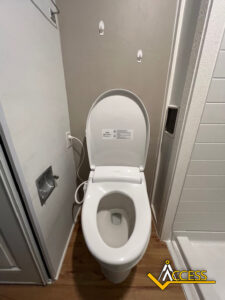 A modern white toilet with the lid open is positioned next to a shower and a wall outlet, on a wooden floor in a bathroom. Two wall hooks and a plumbing access panel are visible nearby.