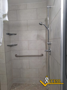 Accessible walk-in shower with beige tiles, a handheld showerhead on a vertical bar, two corner shelves, a horizontal grab bar, and a curtain partially visible on the left. ACCESS logo in bottom right corner.