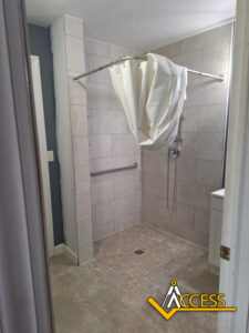 Accessible walk-in shower with beige tiles, a handheld showerhead, a grab bar on the wall, a partial shower curtain gathered at the top, and the Access company logo in the bottom right corner.