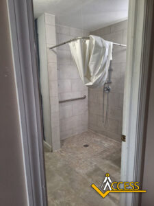 Accessible walk-in shower with tiled walls and floor, equipped with a grab bar, handheld showerhead, and a partially closed shower curtain. The word ACCESS with a yellow triangle logo is visible in the bottom right corner.