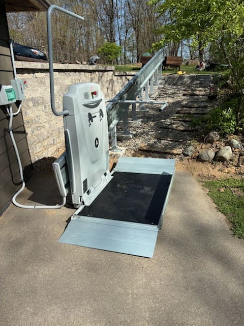 A motorized outdoor wheelchair lift with a ramp is installed on a concrete path, leading up a sloped track beside a landscaped yard with rocks and trees in the background.