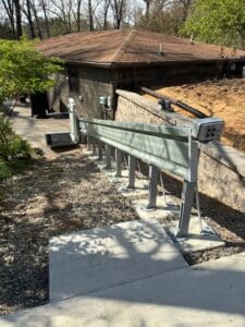 A metal track system is installed alongside a concrete path on a slope, leading down to a house with a brown roof. Trees and greenery surround the area, and the scene is brightly lit by sunlight.