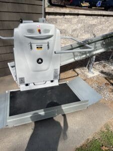 A motorized wheelchair platform lift with safety rails and ramp is installed outdoors next to a building, allowing accessibility up a small flight of stairs. A person’s shadow is visible on the ground in front.