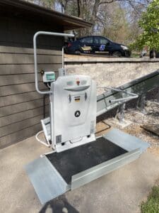 A motorized outdoor wheelchair platform lift is installed by a ramp next to a building, with a safety rail and control panel. In the background, a car with wheelchair icon decals is parked by a stone wall.